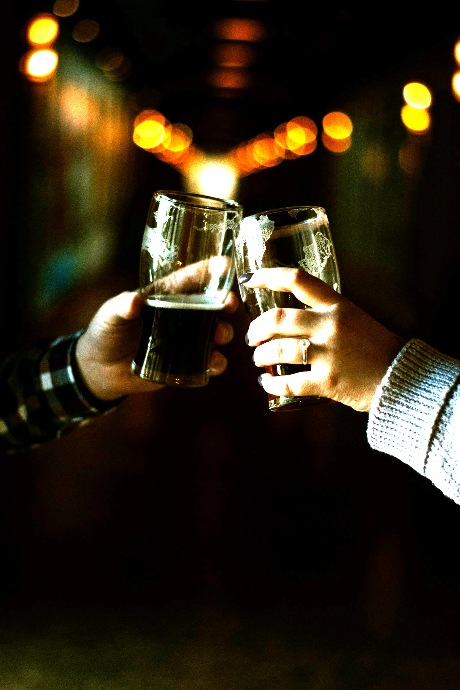 Two beer glasses clinking in a dark bar, warm amber lights behind